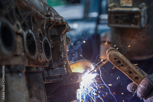 Close-up of a professional welder wearing safety gear, welding a metal car engine part with sparks flying in an industrial workshop.