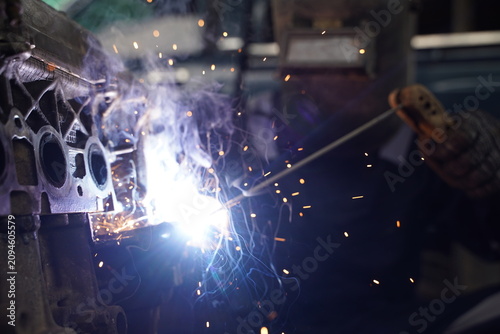 Close-up of a professional welder wearing safety gear, welding a metal car engine part with sparks flying in an industrial workshop.