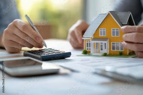 hands calculating home purchase with pen on a calculator beside a yellow model house and paperwork, focused decision-making