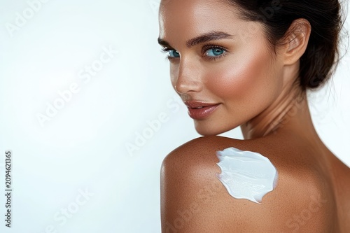 Confident woman with glowing tanned skin and blue-green eyes looking over her shoulder with white moisturizer on her shoulder