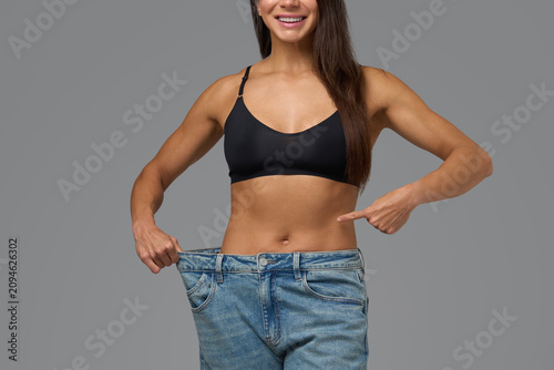 Smiling athletic woman in black sports bra proudly showing loose jeans after fitness transformation