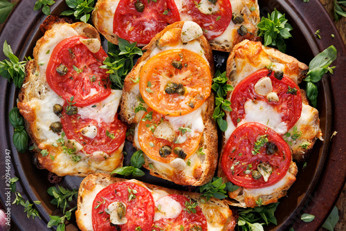 Bruschettas with mozzarella and grana padano cheese, tomatoes, capers and garlic seasoned with aromatic herbs on a plate, top view, close-up view
