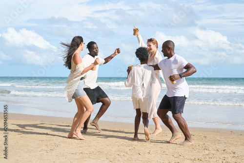 Group of five diverse friends joyfully dancing and celebrating on a sandy beach by the ocean, under a partly cloudy sky