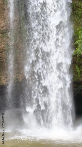 Vertical close up of a powerful waterfall cascading down a rocky cliff, creating misty water spray in a lush green forest.
