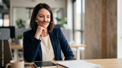 Funny young brunette businesswoman in blue blazer holding pencil with nose and lips at office table. Concept of work procrastination, boredom, corporate humor and creative block.