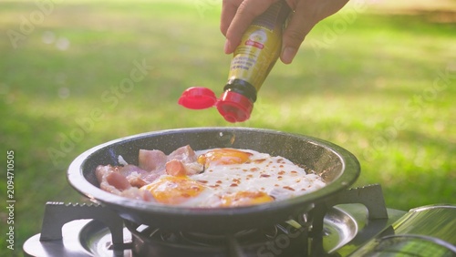 Fotografía A person seasons fried eggs and bacon in a pan on a portable stove, preparing an enjoyable camping breakfast while cooking outdoors on a sunny day in the park