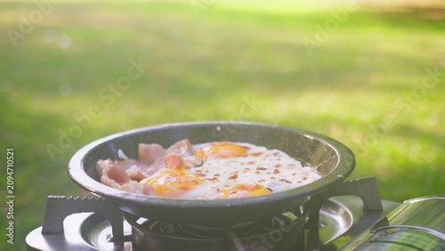 Fotografía A person seasons fried eggs and bacon in a pan on a portable stove, preparing an enjoyable camping breakfast while cooking outdoors on a sunny day in the park