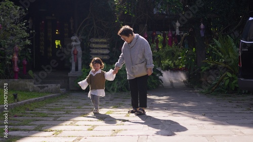 Tableau sur toile A caring Asian grandmother guides her joyful toddler granddaughter along a sunlit garden path, a cherished moment of learning and intergenerational bonding