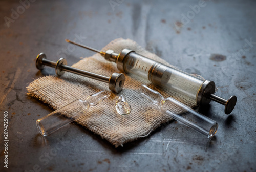 Three-quarter view of a disassembled vintage glass and steel syringe on a crumpled gauze pad. Broken clear glass ampoules rest on a dark patinated steel plate with heat stains.
