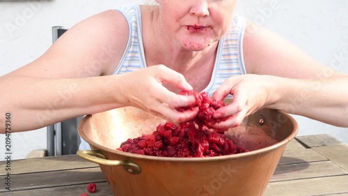 Blonde woman wearing a tank top sits at a wooden table, enthusiastically eating handfuls of raspberries from a large copper basin, her face and hands stained with red juice