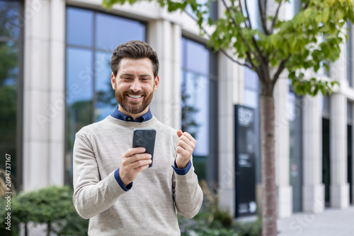 Smiling young man standing on the street near an office building, received a message with better results, rejoices in success