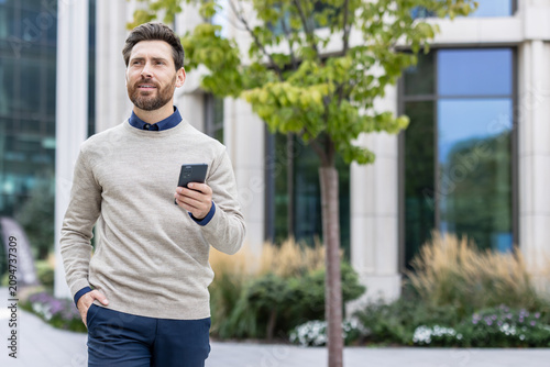 A young businessman is walking down the street near an office center, holding a phone in his hand and looking to the side with a smile
