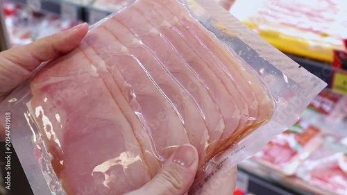 Close up of female hands holding and inspecting plastic vacuum package of sliced ham or pork loin in a supermarket. Customer choosing meat cold cuts in a grocery store. Shopping concept.