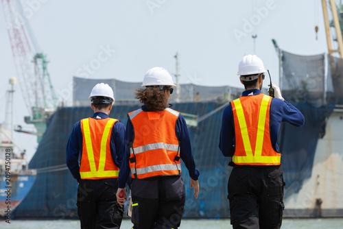 industrial worker walking towards large cargo ship at bustling shipyard wearing white hard hat and bright orange reflective vest feeling determined about upcoming project