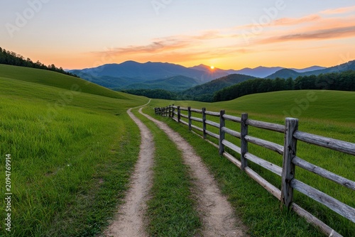 A winding dirt path runs alongside a wooden fence through lush green fields toward distant mountains under a colorful sunset sky.