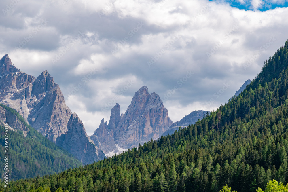 Fototapeta premium Dramatic view of rugged dolomite mountain peaks rising above dense green pine forests under a clouded blue sky, Dolomites, Italy