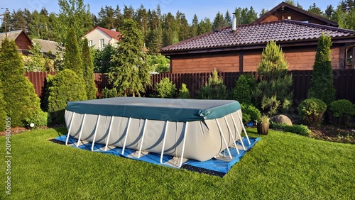 On the mown lawn there is a rectangular frame PVC swimming pool, covered with a cover. Nearby there are thujas, pines, flower beds, fences and buildings stands. Sunny summer weather and blue sky