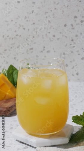 Summer mango drink mocktail with ice and mint on a white coaster, styled in bright sunlight on a terrazzo countertop with fresh mango and clean copy space. A clear rounded glass holds a golden mango