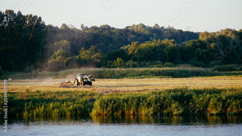 Bilde på lerret A tractor rakes mown grass in a meadow by the river, against the backdrop of a d