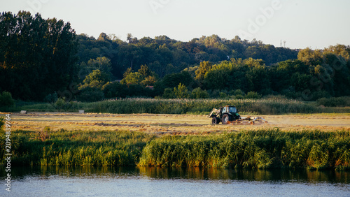 Tableau sur toile A tractor rakes mown grass in a meadow by the river, against the backdrop of a dense forest in the warm rays of the sun