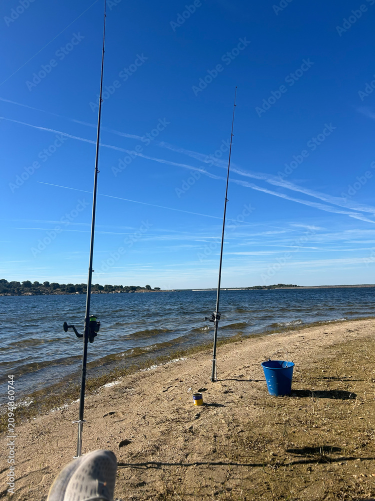 Fototapeta premium Fishing Rods On A Sandy Lake Shore With Bucket And Calm Blue Sky For Relaxing Day