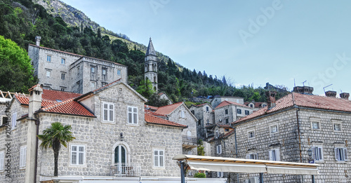 Scenic waterfront view of the historic town of Perast on the Bay of Kotor, Montenegro. The image features the town's distinctive bell tower, traditional stone architecture, small boats, and dramatic m
