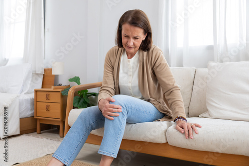 Foto Older woman sitting on a couch holding her knee in pain during morning