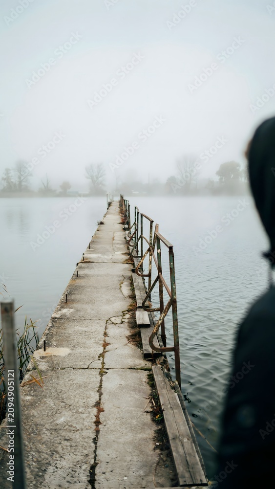 Fototapeta premium Person Standing Near Pier on Calm Lake with Misty Weather and Overcast Sky
