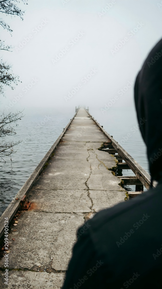 Fototapeta premium Man Standing Near Edge of Pier Over Calm Foggy Lake on a Misty Day