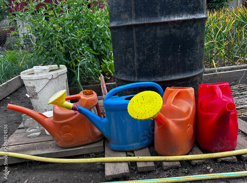 Garden watering cans and plastic containers are arranged near a water barrel. Perfect for horticulture articles, DIY guides, and editorial layouts.