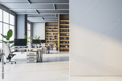 Bright open space office interior with white desks, computer monitors, wooden bookshelves, green plants and empty wall mockup in foreground. 3D Rendering