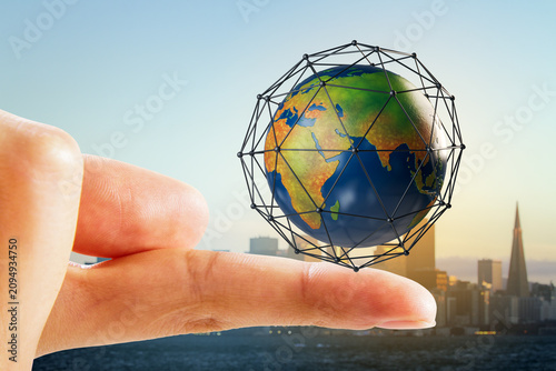 Fotografie Miniature Earth globe in polygonal wireframe sphere balancing on fingertip against blurred city skyline background illustrating global connection network