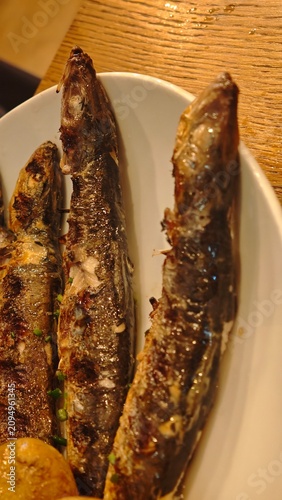 Grilled fish served on a white plate with a side of baked potato and garnished with herbs on a wooden table in a restaurant setting