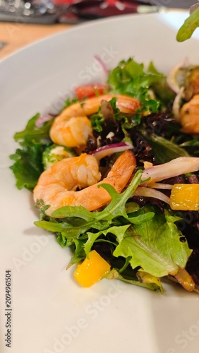Shrimp salad with vibrant mixed greens, red onion, and mango chunks presented in a white bowl on a wooden surface with blurred background details