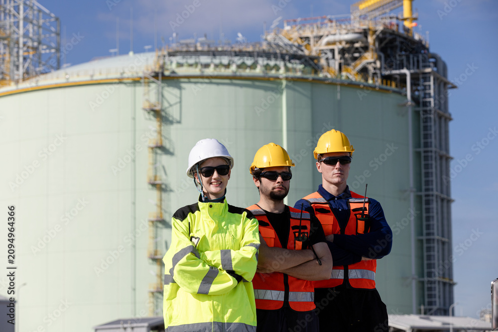 Fototapeta premium Team of engineers and construction workers wearing safety gear collaborating at an industrial site featuring large storage tanks