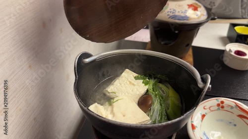 Hand opening a hot nabe pot revealing a steaming traditional Japanese meal with tofu and vegetables