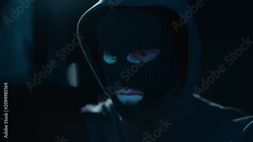 Hacker in balaclava and hood at laptop in dark room