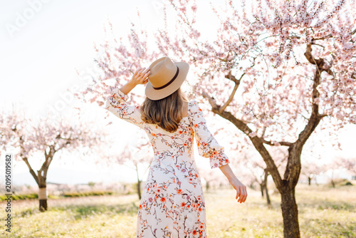 A young woman in a dress and hat walks through a garden surrounded by blossoming trees. A rear view of the woman enjoying nature or almond trees at sunset. Concept of beauty, nature, and relaxation.