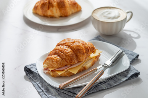 A golden croissant sandwich with ham and cheese on a white plate on a marble background