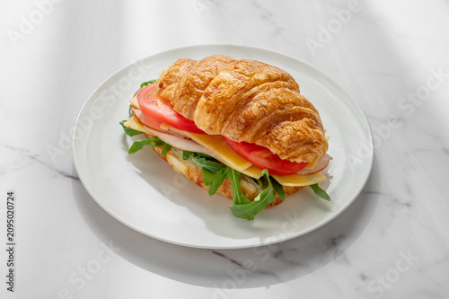A golden croissant sandwich with ham, cheese, tomato, and arugula on a marble background