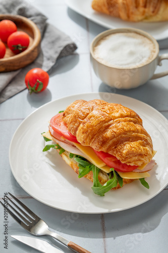 A golden croissant sandwich with ham, cheese, tomato, and arugula on a tile background