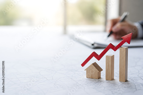House model beside rising wooden bars and a red upward arrow on architectural plans, representing property value growth, real estate investment and housing market trends.