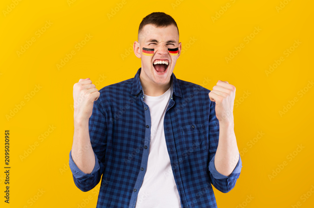 Fototapeta premium Excited Young Caucasian Man With Face Paint Showing Happiness By Raising Fists While Celebrating Sports Event In Bright Yellow Studio Background. Sports Concept.
