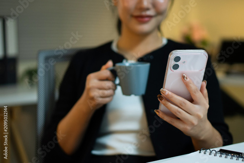 A close-up shot of an Asian businesswoman's hands holding a pink smartphone and a cup of coffee during a late-night office break.