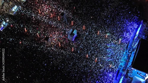 Aerial around view of crowd at night music concert. People gathered at festival outdoor stage with lights and screens, 4k