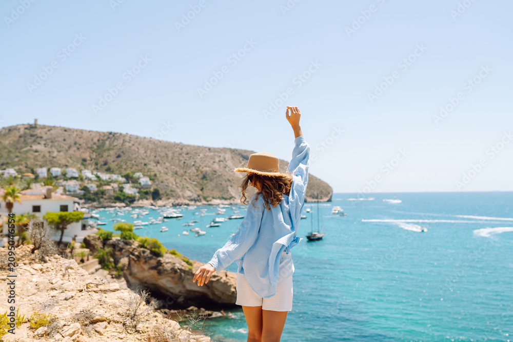 Obraz premium A rear view of a young woman wearing a hat sitting on the edge of a cliff. The woman feels free and enjoys the seascape on a sunny day. Concept of nature, relaxation, and freedom.