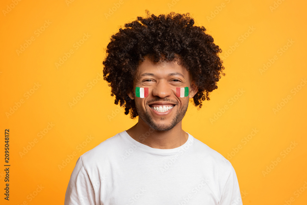 Fototapeta premium Smiling Black Man In His 30s With Italian Flag Face Paint, Enjoying Celebration, Standing Against Bright Yellow Background. Emotion Of Happiness Reflecting Festive Atmosphere, Concept Of Celebration.