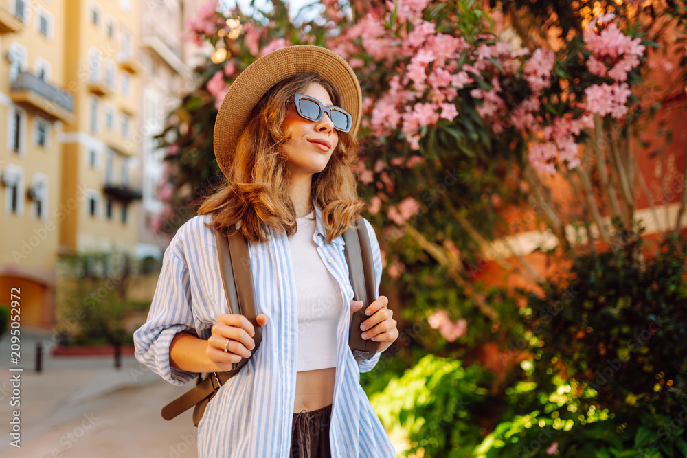 Fototapeta premium Young woman wearing hat and carrying backpack near blossoming trees on sunny day. Woman in sunglasses admires city's architecture. Lifestyle. Concepts of tourism, vacation, and rest.