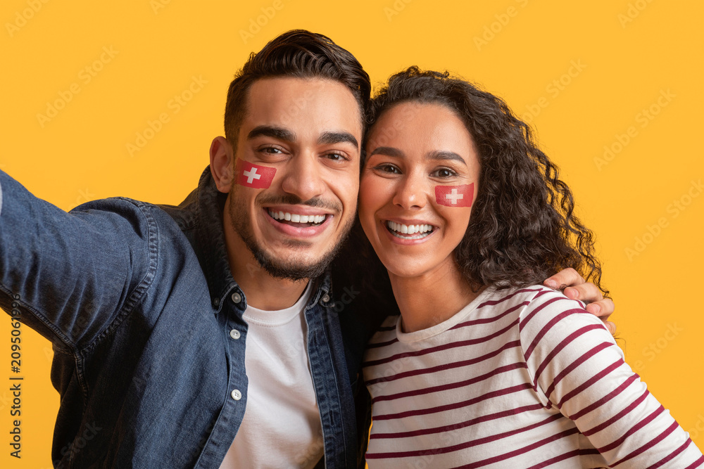 Fototapeta premium Smiling Multiracial Young Couple With Face Paint Celebrating At Studio With Yellow Background. Happy Emotion Displayed While Taking Selfie Together. Joyful Moment To Capture At Party.