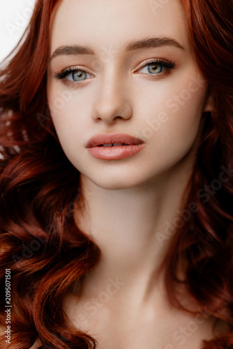 Close-up portrait of a young woman with wavy red hair, blue eyes, and natural makeup against a clean white background, with soft even lighting and a calm